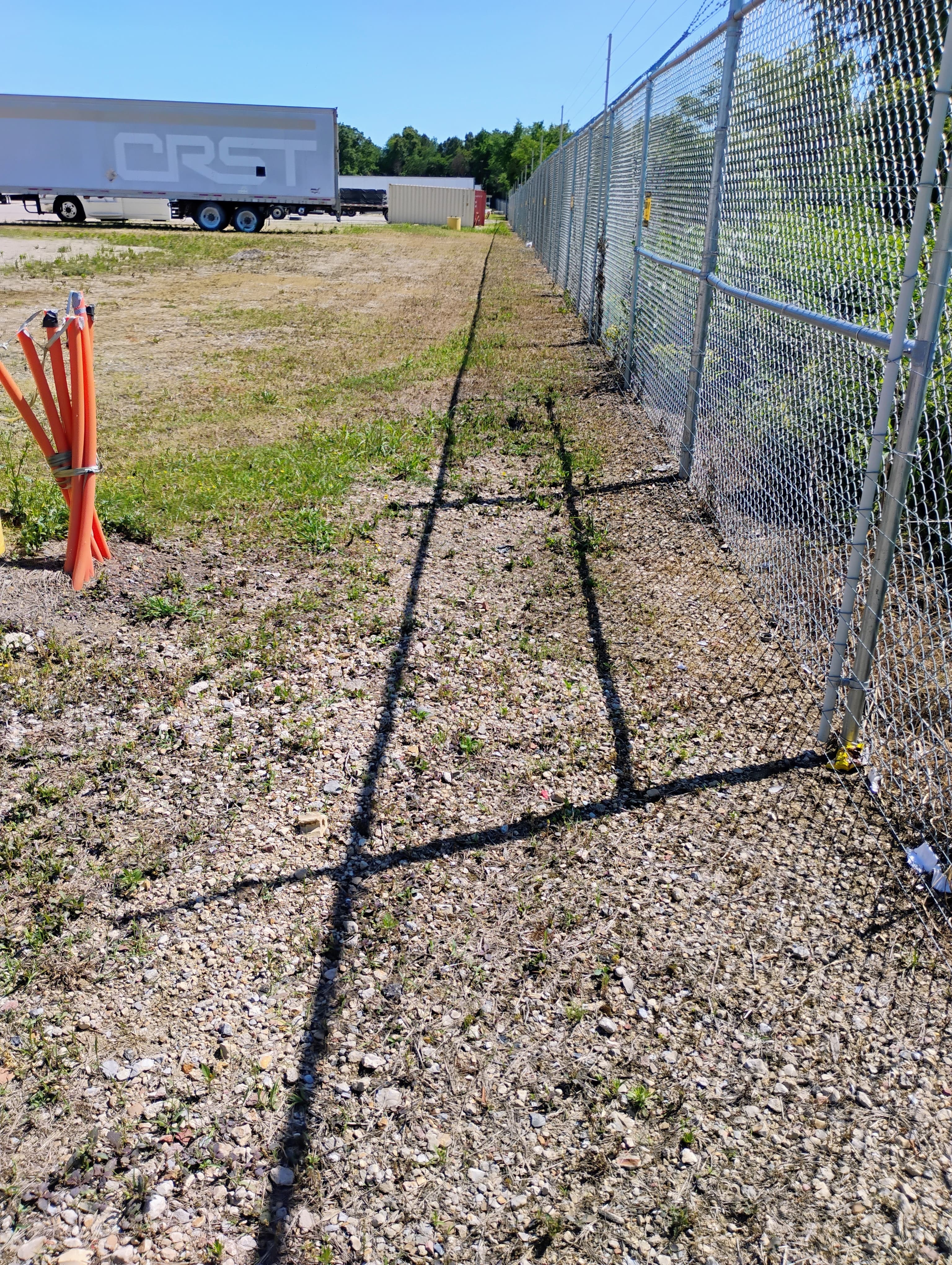 Chain-link fence casting a long shadow on gravel beside a parked CRST trailer.