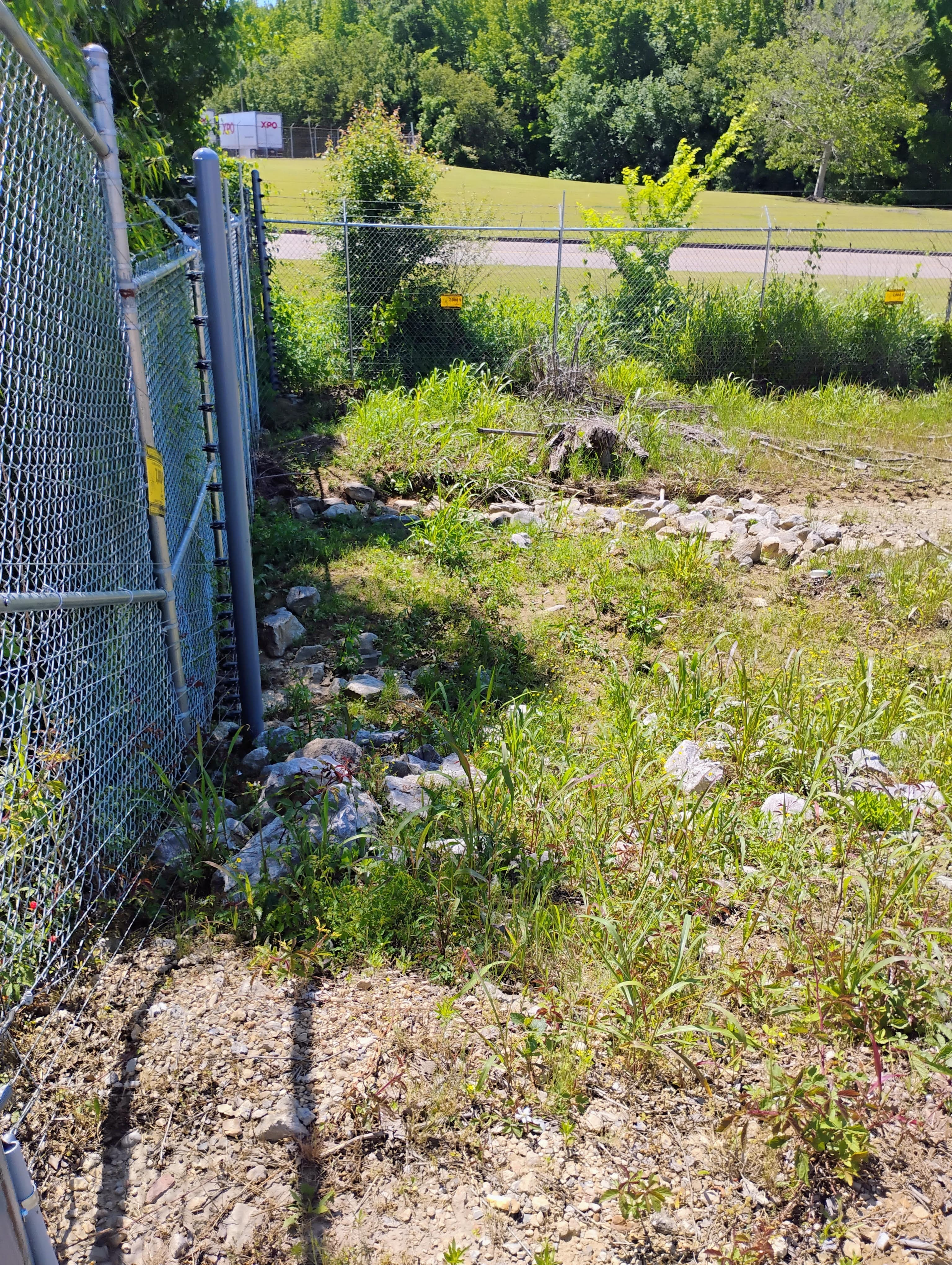 Chain-link fence beside a rocky, grassy area with a white truck and forest background.